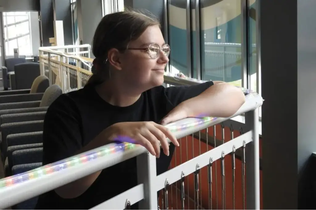 A person wearing glasses and a black shirt leans on a railing wrapped with colorful lights, looking out of large windows in a modern indoor space.